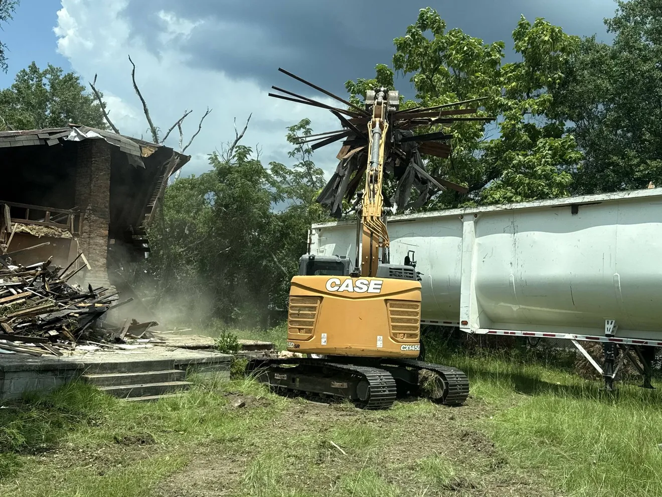 CASE excavator pulling down the roof framing of an old building, debris piled on the slab beside a white dump trailer