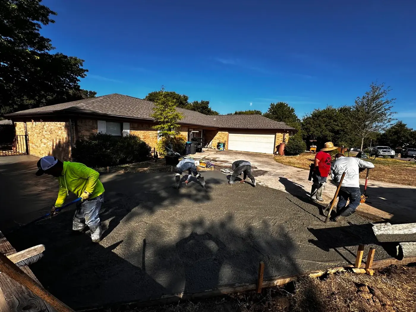 A crew finishes a freshly poured concrete driveway in front of a brick ranch house